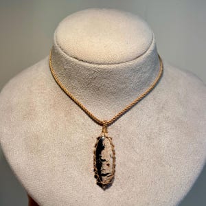 May include: A handcrafted necklace featuring a pendant with a black and cream-colored stone. The stone is encased in a woven, beige-colored cord, suspended from a matching cord necklace. The pendant has an oval shape.