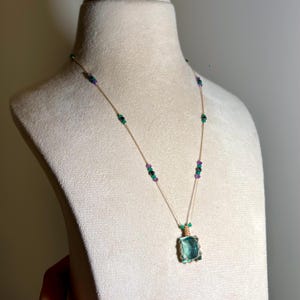 May include: A delicate necklace featuring a rectangular, translucent teal pendant encased in gold-toned wire. The thin chain is accented with small, evenly spaced beads in shades of emerald green, deep purple, and black, creating a subtle contrast.