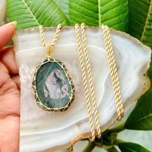 Può includere: Una collana con catena dorata con un ciondolo in pietra verde e rosa. La pietra è avvolta in filo d'oro e ha una forma naturale unica.