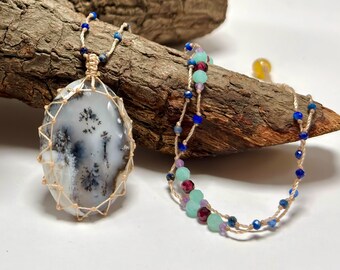 May include: A handcrafted necklace featuring a large, oval-shaped pendant with a dendritic pattern in shades of gray and white, encased in a woven beige cord. A separate beaded necklace with blue, green, and purple beads complements the pendant.