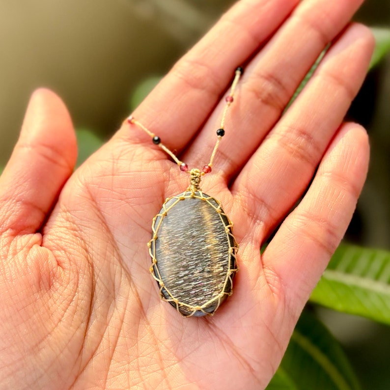 May include: An oval pendant with a black and silver sunstone wrapped in gold wire. The pendant is on a thin gold chain with small red and black beads.