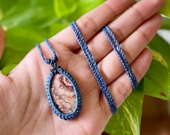 Könnte beinhalten: Eine blaue Makramee-Schnurkette mit einem braunen und weißen Achat-Anhänger. Der Anhänger ist oval geformt und hat ein wirbelndes Muster.