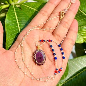 May include: A handmade necklace featuring a large, oval-shaped, deep red gemstone pendant wrapped in gold wire. The necklace has a delicate, beaded chain with white, blue, and red accents. The necklace is displayed against a natural green background.