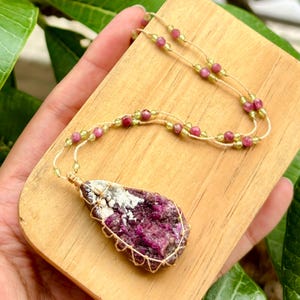 Op de afbeelding: Een roze en groene edelsteen ketting met een grote, ruwe roze edelsteen hanger gewikkeld in gouden draad. De ketting is gemaakt met kleine roze en groene kralen op een natuurlijk vezelkoord.