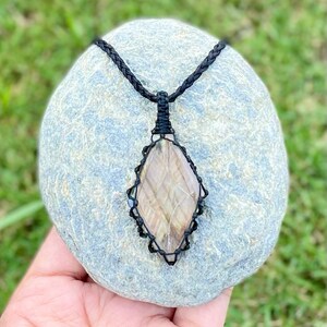 Pode incluir: Um pingente de pedra preciosa marrom e branca com um colar de cord&atilde;o preto. A pedra preciosa est&aacute; engastada em um envolt&oacute;rio de fio preto e tem a forma de um diamante.
