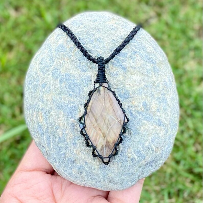 Pode incluir: Um pingente de pedra preciosa marrom e branca com um colar de cord&atilde;o preto. A pedra preciosa est&aacute; engastada em um envolt&oacute;rio de fio preto e tem a forma de um diamante.