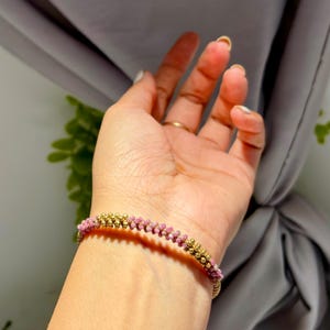 May include: A gold-coloured bracelet featuring alternating rows of small, round, pink beads and gold-coloured beads. The bracelet is worn on a wrist, with a gold ring on a finger. The background is a soft, neutral colour.