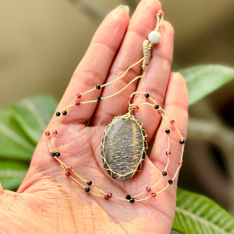 May include: A gold and silver gemstone pendant necklace with black and red beads. The pendant is wrapped in gold wire and hangs from a thin, beige cord.