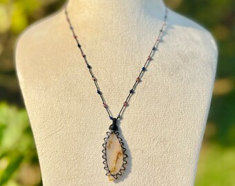 May include: A white agate pendant with brown and black markings is wrapped in black cord and hangs from a black cord necklace with small blue and pink beads.