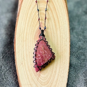May include: A pink and red gemstone pendant necklace with black cord and beads. The pendant is a triangular shape with a rough, natural texture.