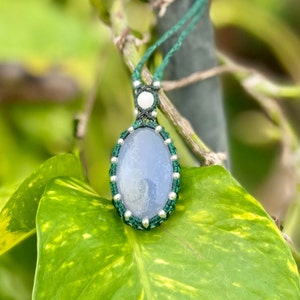 May include: A handcrafted necklace featuring a large, oval, pale blue gemstone pendant. The stone is encased in a macrame design with green and silver accents, suspended from a green cord. The background is blurred, with green foliage.