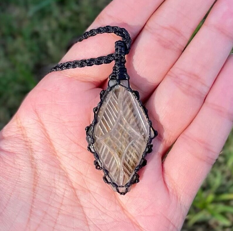 Pode incluir: Um pingente de cristal de quartzo transparente com um design geom&eacute;trico, envolto em um cord&atilde;o de macram&ecirc; preto.