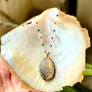 May include: A gold necklace with a large, oval-shaped sunstone pendant wrapped in wire. The necklace is made with black and red beads.