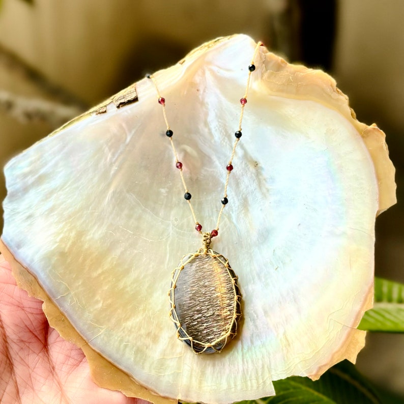 May include: A gold necklace with a large, oval-shaped sunstone pendant wrapped in wire. The necklace is made with black and red beads.