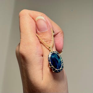 Puede incluir: Un collar con un colgante de piedra preciosa azul oscuro de forma ovalada, encerrado en una delicada jaula de alambre de tono dorado. El colgante cuelga de una cadena de cuerda a juego en tono dorado, con un acabado pulido y un tono azul.