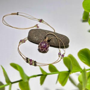 Può includere: Una collana fatta a mano con un grande pendente rotondo di pietra preziosa viola scuro, racchiuso in un cordoncino beige intrecciato. La collana ha un design delicato e stratificato con piccole perline colorate. Lo sfondo è bianco neutro con una pietra naturale.