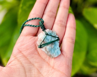 Pode incluir: Um cordão verde envolto em torno de um pingente de cristal azul claro e branco. O pingente está amarrado a um cordão verde.
