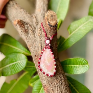 May include: A pink and white gemstone pendant necklace with a red cord and silver beads. The pendant is a teardrop shape and is surrounded by silver beads.
