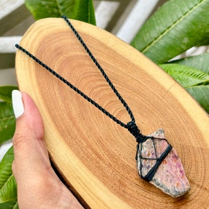 May include: A pink and white gemstone pendant necklace with a black cord. The gemstone is wrapped in black cord and has a rough, natural shape.