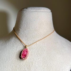 May include: A necklace featuring a polished, oval-shaped pink and white stone pendant encased in a gold-colored wire cage. The pendant hangs from a matching gold-colored twisted cord chain, creating a simple and cohesive jewelry piece.
