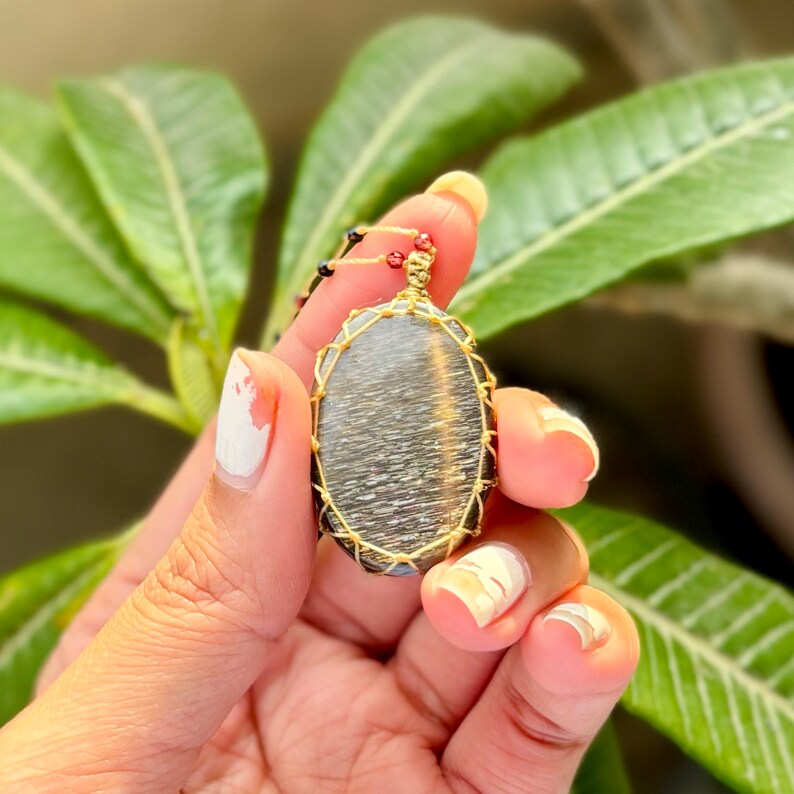 May include: An oval pendant with a black and gold gemstone, wrapped in gold wire. The pendant is hanging from a black cord with red beads.