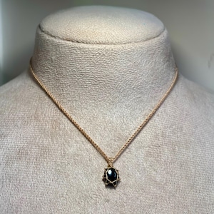 May include: A gold-colored necklace with a black oval pendant. The pendant is encased in a gold-colored wire cage. The necklace is displayed on a mannequin.