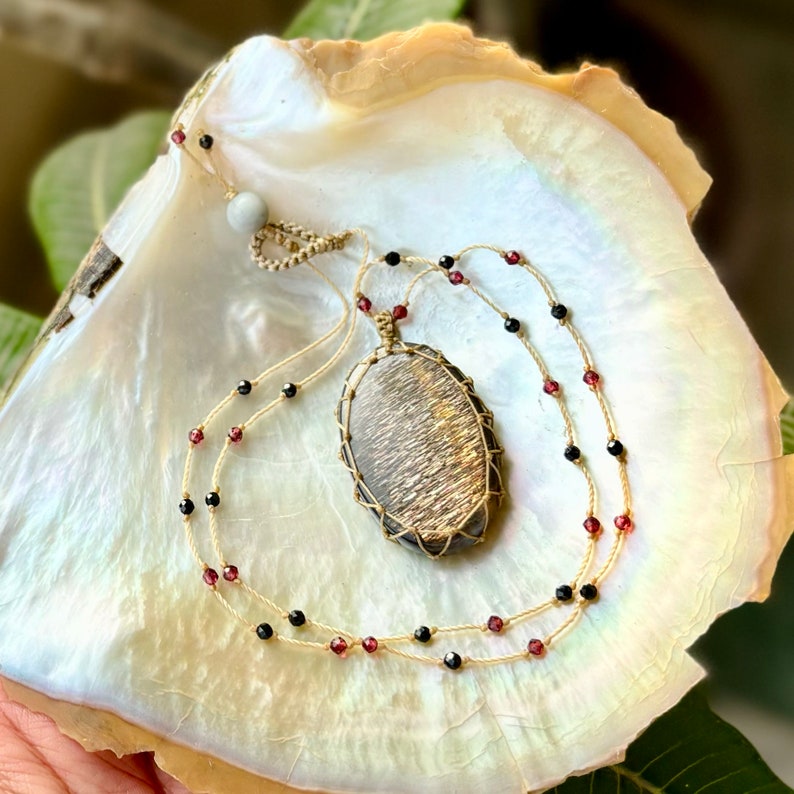 May include: A gold and brown stone pendant necklace with black and red beads on a beige cord. The necklace is displayed on a white and beige shell.