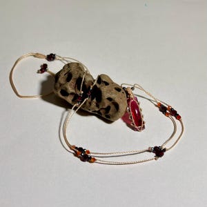 May include: A necklace featuring a translucent red cylindrical pendant encased in a tan cord macrame cage. The cord is adorned with small dark red and orange beads, and the necklace is draped over a porous, light brown, irregularly shaped stone.