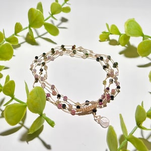 Op de afbeelding: Een meerstrengs armband met roze, groene en zwarte kralen op een beige koord. Een druppelvormige roze bedel hangt aan de armband. De armband is omgeven door groen gebladerte. De kralen zijn waarschijnlijk ongeveer 0,5 cm in diameter.