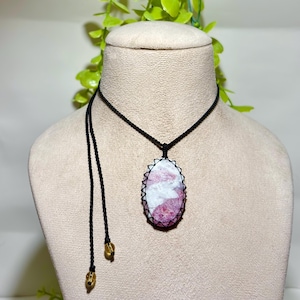 May include: A handmade necklace featuring a pink and white oval gemstone pendant. The pendant is encased in a macrame design and hangs from a black cord. The necklace has a long, adjustable cord with gold-colored beads.