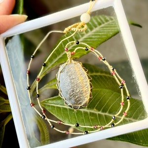 May include: A gold wire wrapped pendant featuring a black and gold gemstone. The pendant is on a beige cord necklace with black and red beads.