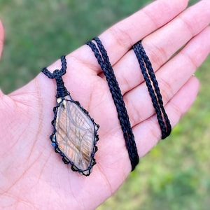Pode incluir: Um pingente de labradorita marrom e dourado com um colar de cord&atilde;o de macram&ecirc; preto. O pingente tem a forma de um diamante e apresenta um design tecido.
