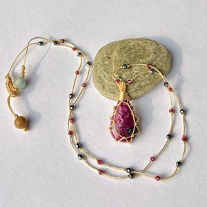 May include: A pink ruby gemstone pendant necklace with gold wire wrapping. The necklace is made with a beige cord and has small black and red beads.