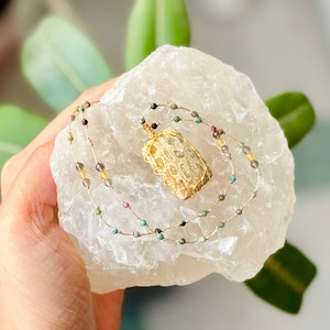 May include: A handcrafted necklace featuring a rectangular, light brown pendant wrapped in gold wire. The necklace has a delicate chain adorned with small, colorful beads in shades of green, pink, yellow, and black. The necklace is displayed on a white, textured surface.