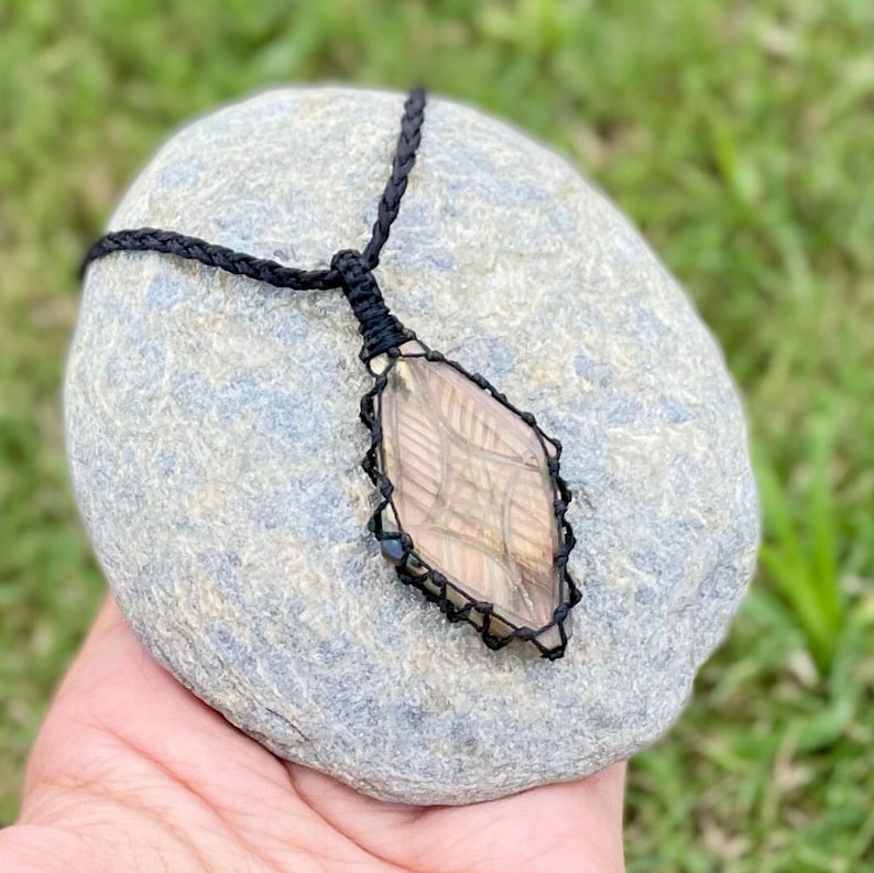 Pode incluir: Um pingente de pedra preciosa marrom e preto com formato de diamante, envolto em um cord&atilde;o preto e colocado em uma rocha cinza.