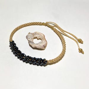 Può includere: Un braccialetto intrecciato beige con una sezione di perline nere. Il braccialetto ha cordoncini beige regolabili con perline color oro alle estremità. Accanto al braccialetto c'è una piccola conchiglia di colore chiaro.