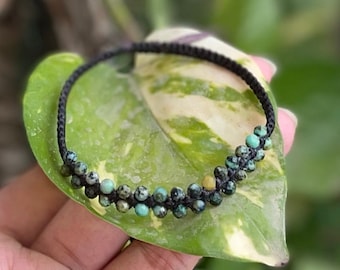 Pode incluir: Um bracelete de cordão preto com contas turquesa e verdes. O bracelete está sobre uma folha verde.