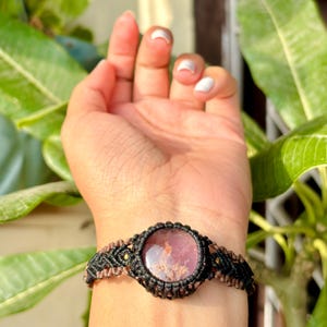 May include: A black and brown macrame bracelet with a pink and white gemstone in the center.