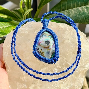 Può includere: Una collana in cordoncino di macramè blu con un grande ciondolo in madreperla di abalone iridescente. Il ciondolo ha una forma a goccia e ha un aspetto naturale e organico.