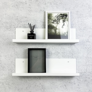 May include: Two white floating shelves mounted on a textured wall. The top shelf holds a framed picture of a forest scene, a black diffuser bottle, and reeds. The bottom shelf has a framed picture with text.