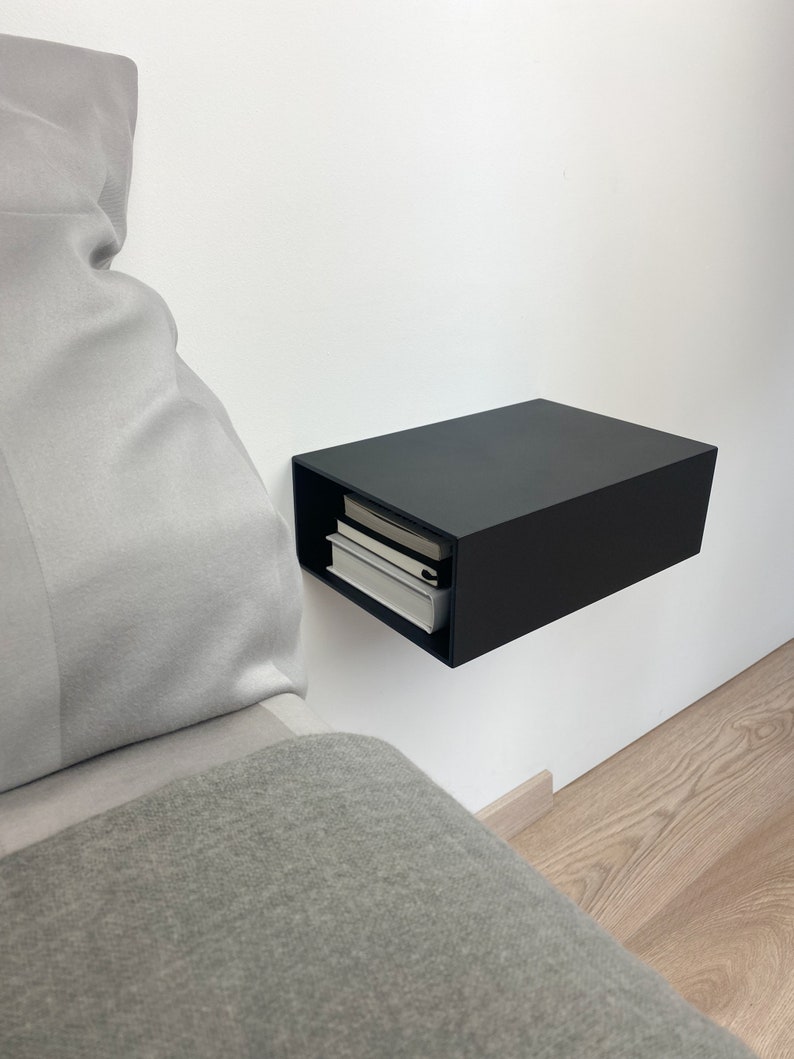 May include: A black floating nightstand with a simple design, mounted to a white wall. The nightstand has a shelf inside and is holding a stack of books. The nightstand is next to a bed with a gray blanket and pillow.