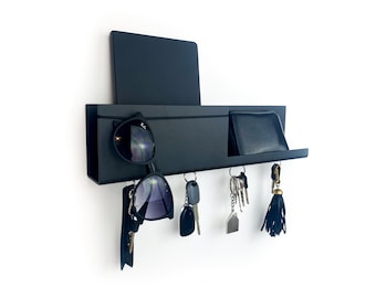 May include: Black wall-mounted key holder with a shelf for sunglasses and a small pocket for a wallet. The key holder has five hooks for hanging keys.