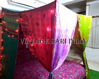 May include: A canopy bed draped with colorful vintage saris, featuring a pink, green, and purple design. The bed is decorated with string lights, creating a warm and inviting atmosphere. The text "Vintage Sari India" is visible in the image.