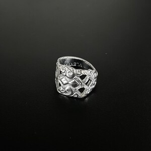 Op de afbeelding: Een zilveren ring met een gedetailleerd, ingewikkeld ontwerp. De ring is breed en heeft een geometrisch patroon met een verhoogd midden.