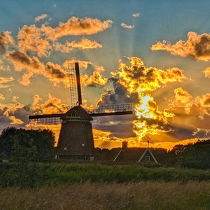May include: A traditional windmill silhouetted against a vibrant sunset sky. The sky is filled with fluffy clouds, some of which are illuminated with a golden glow. The windmill has wooden blades and a brick base.