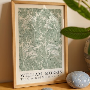 Op de afbeelding: Een ingelijste print met een groen bloemmotief op een crèmekleurige achtergrond. De print zit in een licht houten frame en bevat de tekst "WILLIAM MORRIS The Cleveland Museum of Art" onderaan. De print wordt op een houten oppervlak weergegeven.