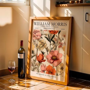 Könnte beinhalten: Ein gerahmter William Morris-Blumendruck lehnt an einer Wand neben einer Weinflasche und einem Glas Rotwein. Der Druck zeigt einen cremefarbenen Hintergrund mit rosa, orangefarbenen und gelben Blumen. Die Weinflasche hat ein rotes Etikett.
