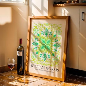 Op de afbeelding: Een ingelijste print met een groen en geel bloemenontwerp, leunend tegen een muur. Een fles rode wijn en een wijnglas met rode wijn staan voor de print. De print luidt "WILLIAM MORRIS CLASSIC TEXTILE PATTERN DESIGN."