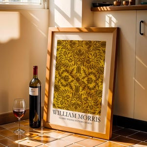 Peut inclure: Une affiche encadrée présentant un motif textile classique de William Morris, dans des tons or et marron. Une bouteille de vin rouge et un verre de vin rouge sont à côté de l'affiche. L'affiche est appuyée contre un mur.