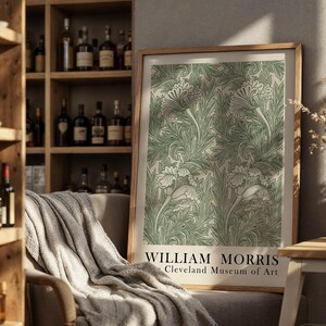 May include: A framed art print featuring a green floral pattern on a cream background. The print, with the text "WILLIAM MORRIS Cleveland Museum of Art," is displayed on a chair with a gray blanket. The room includes a wooden shelf with bottles and a vase with dried flowers.